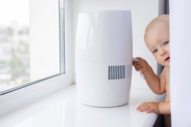 Küçük çocuk perdelerden dışarı bakıyor ve hava nemlendiricisinin güç kablosuna dokunuyor. Çocuk evde dalga geçiyor ve sorun çıkarıyor..