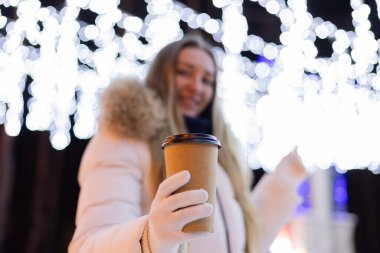 Gülümseyen kız sıcak içecekle bir fincan teklif ediyor. Noel Aydınlatma arka planında. Seçici odak