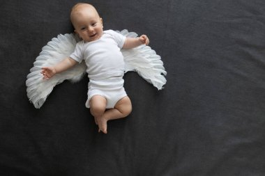 Beyaz giysiler içinde gülümseyen yeni doğmuş bir bebek ve gri bir battaniyenin üzerinde uzanan melek kanatlarıyla