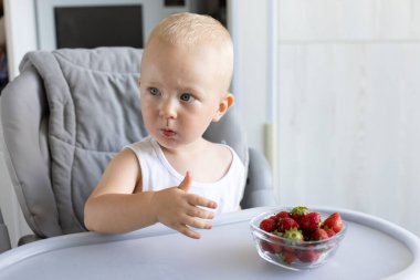 Küçük çocuk yüksek sandalyede oturmuş çilek yiyor.