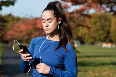 Spor kadını kulaklıkla müzik dinlemek için akıllı telefon kullanıyor. Güneşli bir sonbahar gününde Londra 'da bir parkta..
