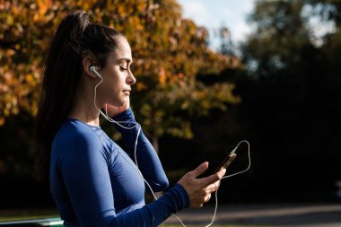 Bir fitness kadını akıllı telefon kullanarak kulaklıkla müzik dinliyor. Bir Londra parkında güneşin tadını çıkarıyor..