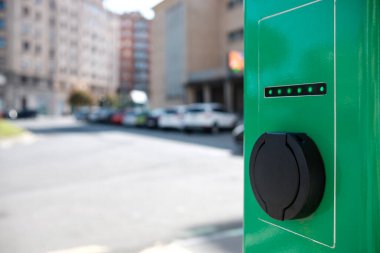 Güneşli bir günde elektrik kartı şarj istasyonu. Bir şehrin ön planı ve odaklanmamış arka planı. Ekolojik kavram.