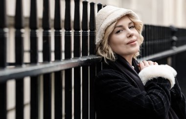 Barlara yaslanıp kameraya bakan genç sarışın kadın portresi. Beyaz şapka, siyah ceket ve eşarp giyiyor..