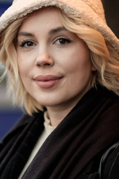 Head-shot of young blonde woman with artificial light. She is looking at camera and she is wearing coat, scarf and cap.