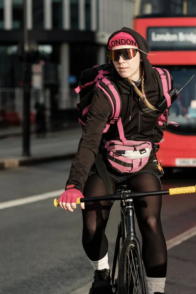 Female bike courier riding her bike with a hand in front of red bus in a London road. She is wearing a cycling outfit. Dangerous job concept.