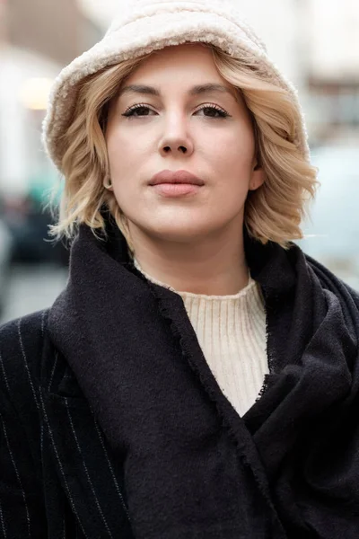 Head-shot of young blonde woman looking at camera. She is wearing a white hat and black coat and scarf.