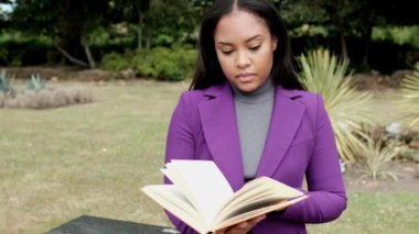 A young woman is reading a book sitting on a stone bench in a London park. She is passing pages and she looks relaxed.