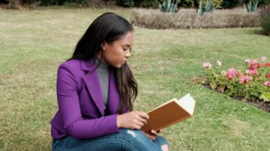 Young woman reading a book sitting on the grass in a London park. There are some beautiful flowers beside her.