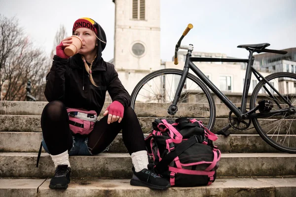 Portrait of bike courier woman taking a break sitting on stairs and drinking a coffee. We can see her backpack and bike on the stairs. An old church behind her. Job break concept.