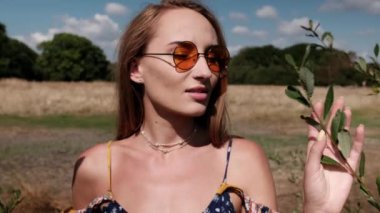 Young woman touching and smelling leaves of a bush. She is wearing sunglasses and a spring dress.