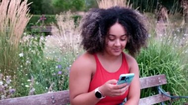 Kıvrımlı genç kadın parkta telefonunu kullanıyor. Mesaj atıyor, bankta oturuyor ve saçları kıvırcık..