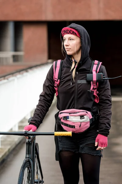 Bike courier woman walking and pushing her bicycle in a London bridge. She is wearing cycling outfit and a pink sling bag.