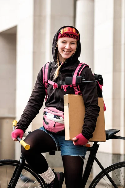 Friendly bike courier woman is sitting on her bike looking at camera. Fashionable cycling outfit. Sling bag. She is holding a parcel. Green-no fumes job concept.