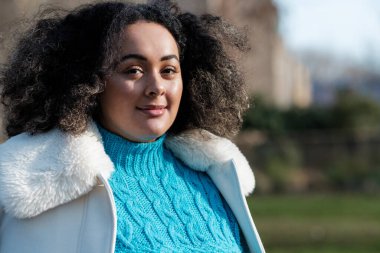 Portrait of smiling young curvy woman looking at camera outdoors in a sunny winter day. She is in a park and wearing white coat and blue sweater.