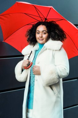 Portrait of curvy woman posing with red umbrella against black wall in London. She is wearing winter outfit. Looking at camera.