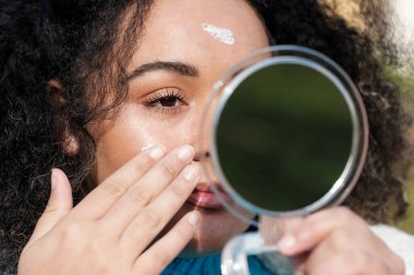 Close-up of young woman applying moisture cream on face outdoors. She is in front of a small mirror and she is using her right hand to use the cream. Skin care and pollution concept.
