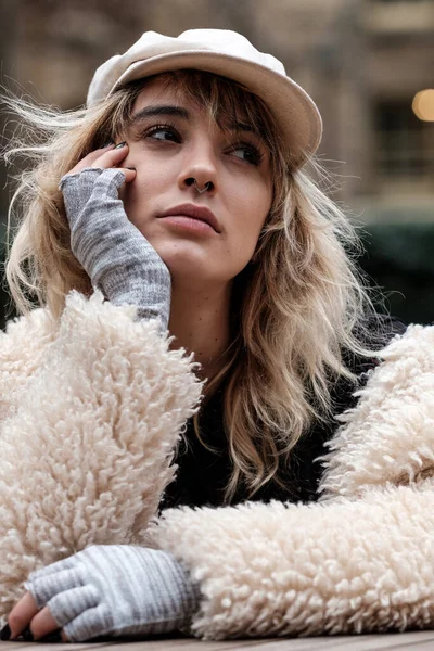 Head-shot of edgy model woman wearing alternative outfit outdoors. She looks pensive and looking away. She is wearing cap, coat and half fingers gloves.