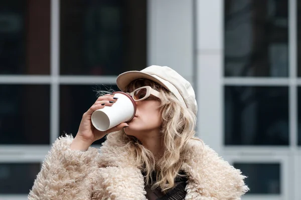 Alternative woman model is drinking coffee outside, in a London street. It is a cold winter day and she is wearing a heavy coat, cap and sunglasses.