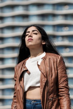 Kentsel bir ortamda güneşin tadını çıkaran rahat Hintli kadın. Arkasında odaklanamadığı yüksek bir bina var..