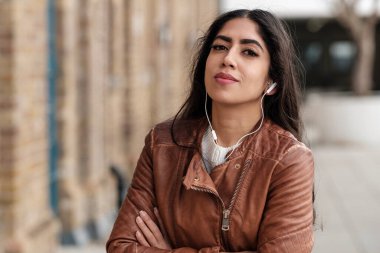 Kendine güvenen Hintli kadın kameraya bakıyor ve müzik dinliyor. Beyaz kulaklık kullanıyor ve deri ceket giyiyor..