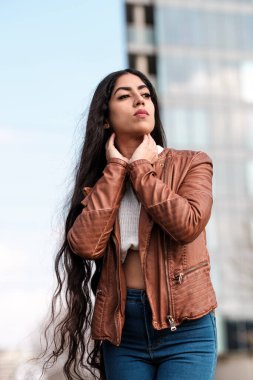 Yüksek binanın önünde poz veren Hintli kadın model. Çok uzun saçları var ve deri ceket giyiyor..