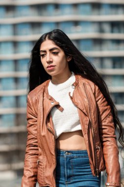 Arkadaki yüksek binayla poz veren Hintli kadın model. Deri bir ceket ve beyaz bir kazak giyiyor. Onun çok uzun saçları var..