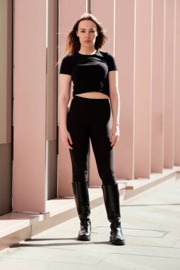 Kafkas modelinin pembe sütunlu tam boy portresi. Siyah bir kıyafet, tişört ve tayt giyiyor..