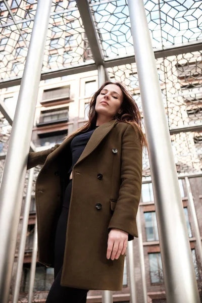 Caucasian woman grabbing a metal tube from structure in street. She is posing with coat and scarf in front of high buildings.