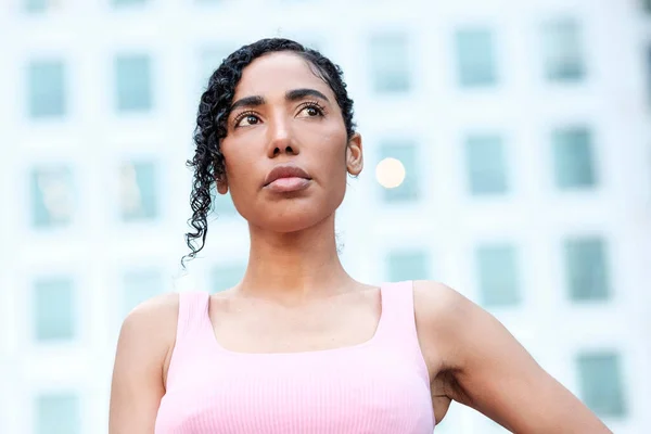 stock image Headshot of young woman showing strength and confidence. There is a blurry building behind her with a lot of windows. Her curly hair is collected and she is wearing a pink sport bra.