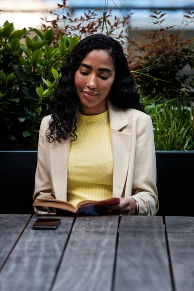 Relaxed woman is reading a book outdoors on a table. She is wearing jacket and yellow t-shirt. There is a phone on a table. Old school hobbies concept.