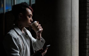 Çift cinsiyetli olmayan erkeğin telefon kullanması ve kahve içmesi. Karanlık ve sade bir hava..