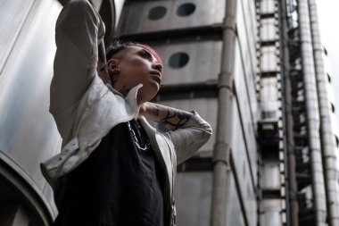 Çift cinsiyetli, çift cinsiyetli olmayan erkek portresi. Gelecekçi ortamda elleri boynun arkasında. Dalgın görünüyor, deri ceket, zincir tasma ve dövmeler. Kimlik, gurur, teğmen ve yaşam tarzı konsepti.