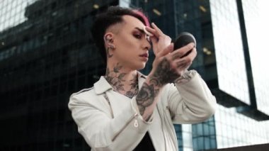 Androgynous erkek dışarıda makyaj yapıyor. Camdan bir binanın önünde ve beyaz bir deri ceket giyiyor. Bir sürü dövme..
