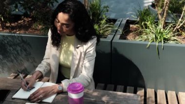 Not defterine yazı yazan çekici bir kadın. Açık havada, güneşli bir günde, masada bir fincan kahve.