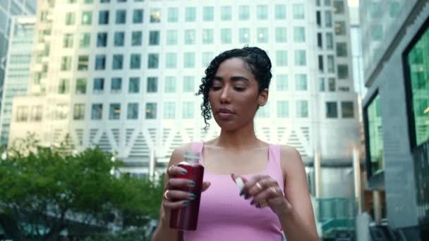 一个年轻健康的女人喝着瓶子里的能量饮料的画像 她身后有一座现代建筑 穿着粉色运动胸罩 — 图库视频影像