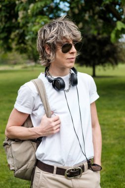 Londra parkında duran çift cinsiyetli bir kadının portresi. Lgbtq ve çeşitlilik konsepti. Boynunda güneş gözlüğü ve kulaklık var. Sırt çantası ve beyaz tişört..