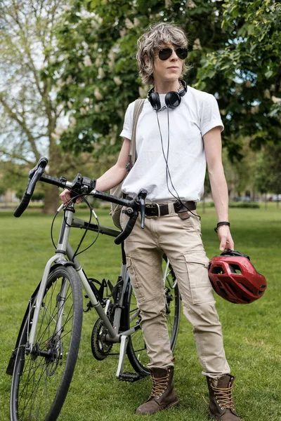 Portrait of androgynous person standing with bike beside. She is in a London park in a cloudy park and she is wearing headphones, sunglasses and red helmet.