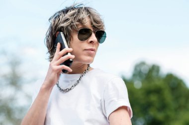 Parkta telefonla konuşan ikili olmayan birinin portresi. Çift cinsiyetli bir görünüşü var ve güneş gözlüğü ve zincir tasma takıyor. Gurur, çeşitlilik ve alternatif yaşam tarzı kavramı.