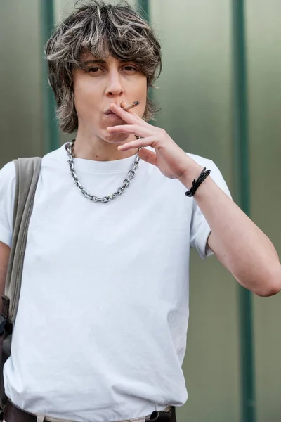 Androgynous non-binary person is smoking a cigarette looking at camera. She is outdoors, a green background behind. Addition and unhealthy habit concept.