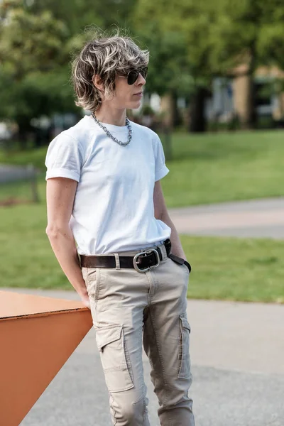Relaxed androgynous person is leaning on an orange table in a London park. Casual outfit, white t-shirt, chain collar and cargo pants.