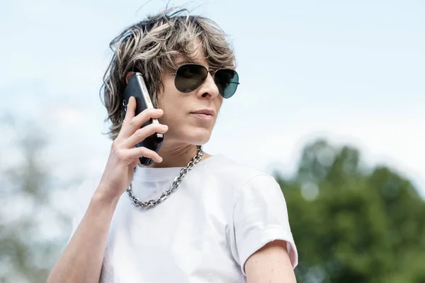 Portrait of non-binary person talking by phone in a park. She has an androgynous looks and is wearing sunglasses and a chain collar. Pride, diversity and alternative lifestyle concept.