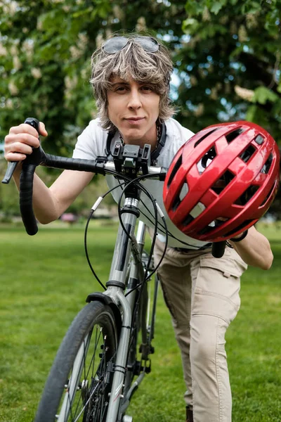 Androgynous non-binary person with a bike in a London park. We can see a red helmet and she is looking at camera. Safety and commute concept.