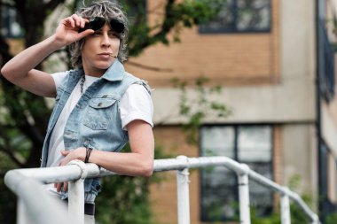 Çift cinsiyetli olmayan Androgynous kadın trabzan köprüsüne yaslanıyor. Güneş gözlüğü ve kot yelek giyiyor..