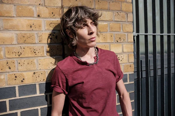 Androgynous non-binary person leaning against brick wall. It is a sunny day and the light is harsh. There is a window protected by bars.