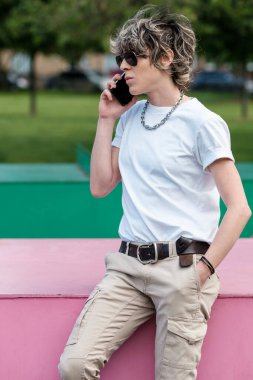 Androgynous ikili olmayan kişi dışarıda telefonla konuşuyor. Parktaki pembe metal bir masaya yaslanıyor..