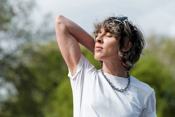 Calm androgynous non-binary person enjoying sunlight in a park. Her hand is behind her neck and she looks happy.