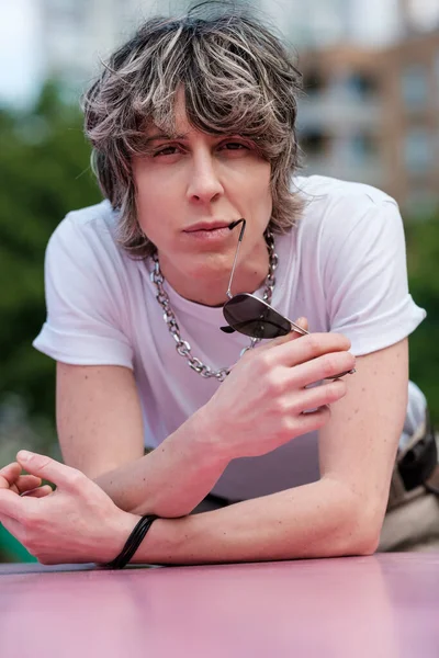 Funny portrait of androgynous person looking at camera outdoors. She is biting the sunglasses temple and she is in a park.