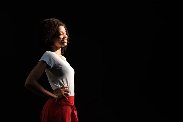 stock image Side view of smiling black woman on black wall. It is summer and she is under harsh sun wearing whiter t-shirt and red skirt.