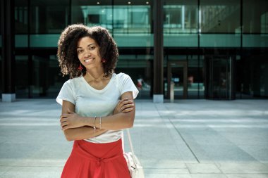 Kozmopolit bir ortamda gülümseyen kadın kıvırcık model. Etrafı modern cam binalarla çevrili ve daha beyaz bir tişört ve kırmızı etek giyiyor..
